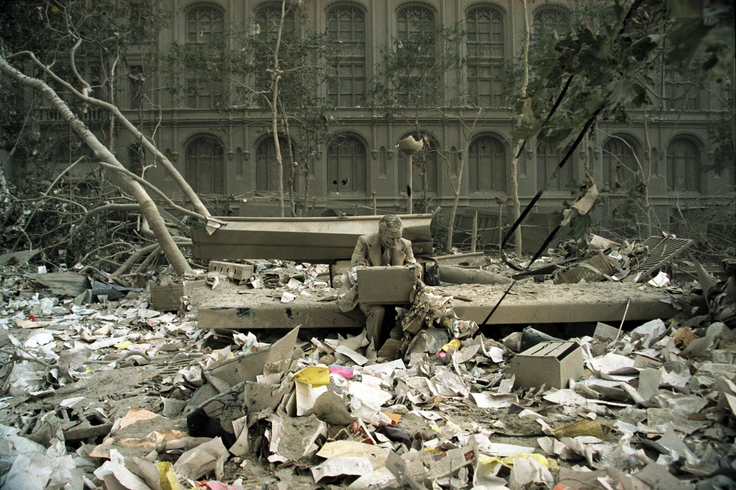 Photograph of a  businessman sitting surrounded by ash and debris, looking inside a ash-coated briefcase. 
