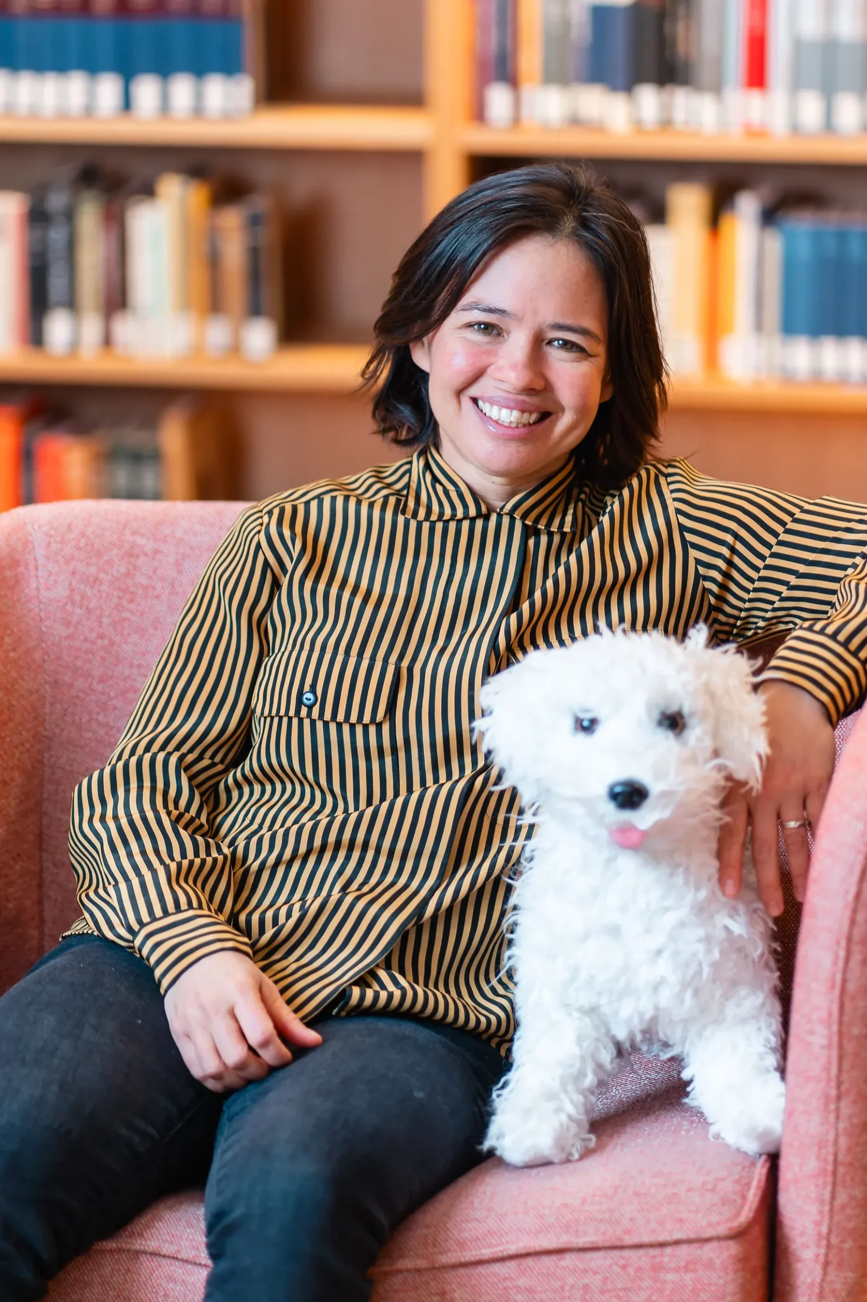 Photograph of Melissa Parrish in a striped button up shirt, sitting on a pink chair next to a stuffed animal version of her dog, "Waldo."