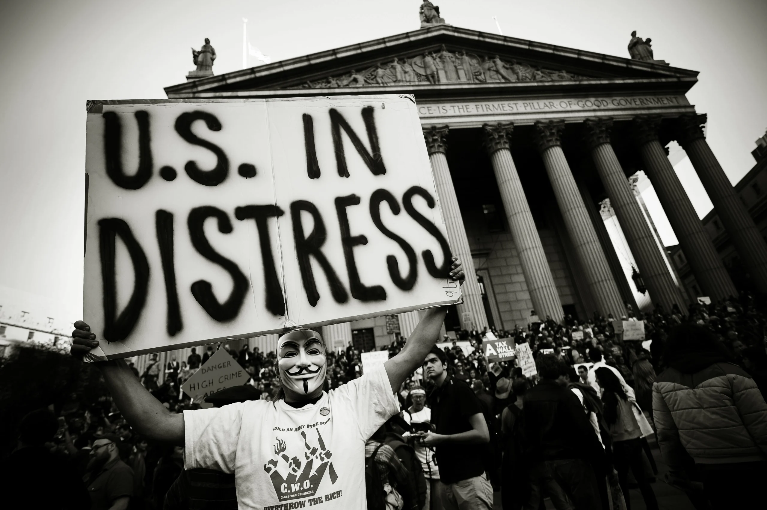 Grayscale photograph of a person in a vendetta mask, holding a sign that reads "U.S. IN DISTRESS" in front of the New York State Courthouse. 