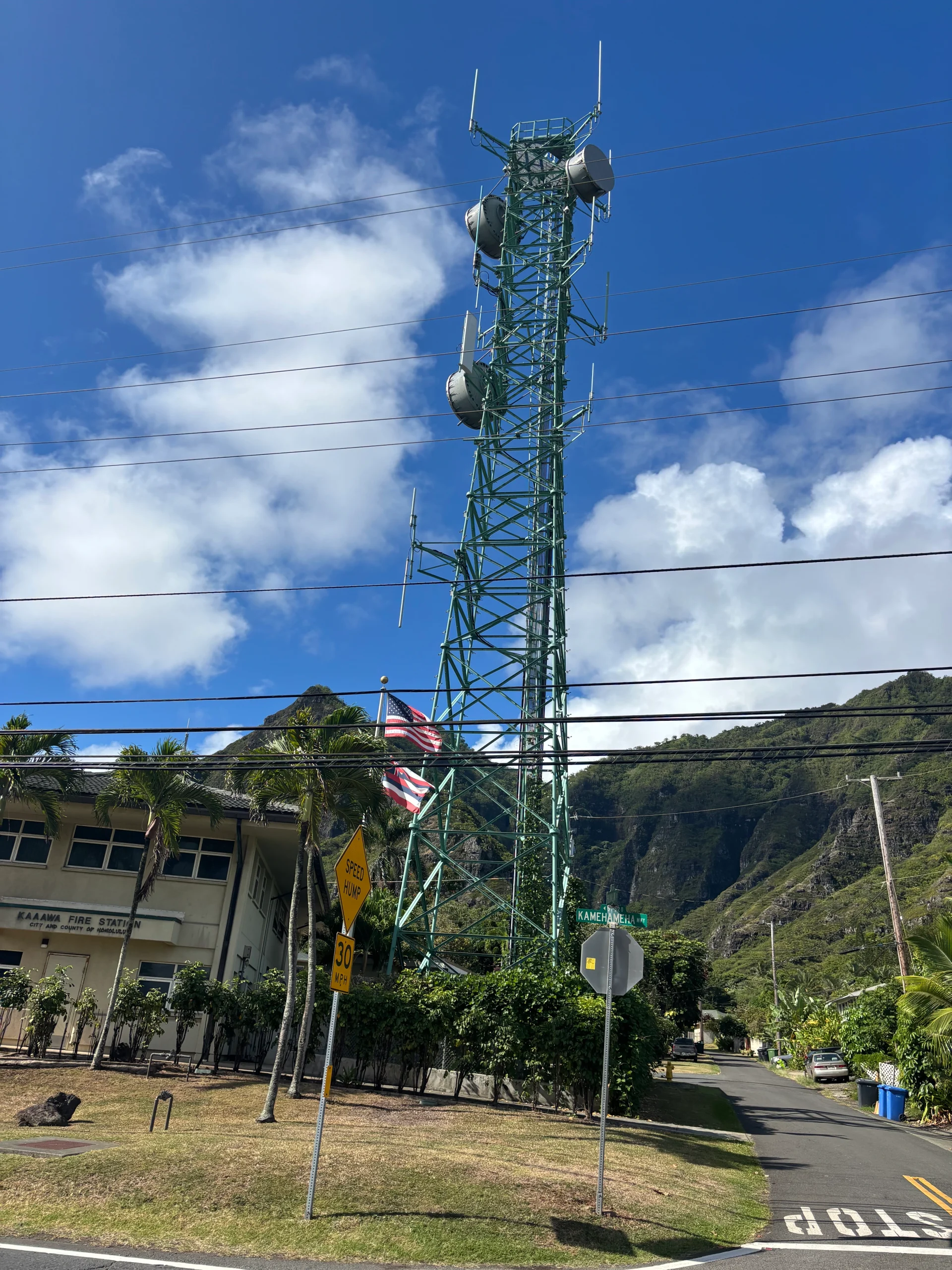 A tall, telephone pole-like structure with antennas extends into a blue sky in front of a flag pole that waves the Hawaiian and U.S. flags. In the background, there are green mountains and palm trees. 