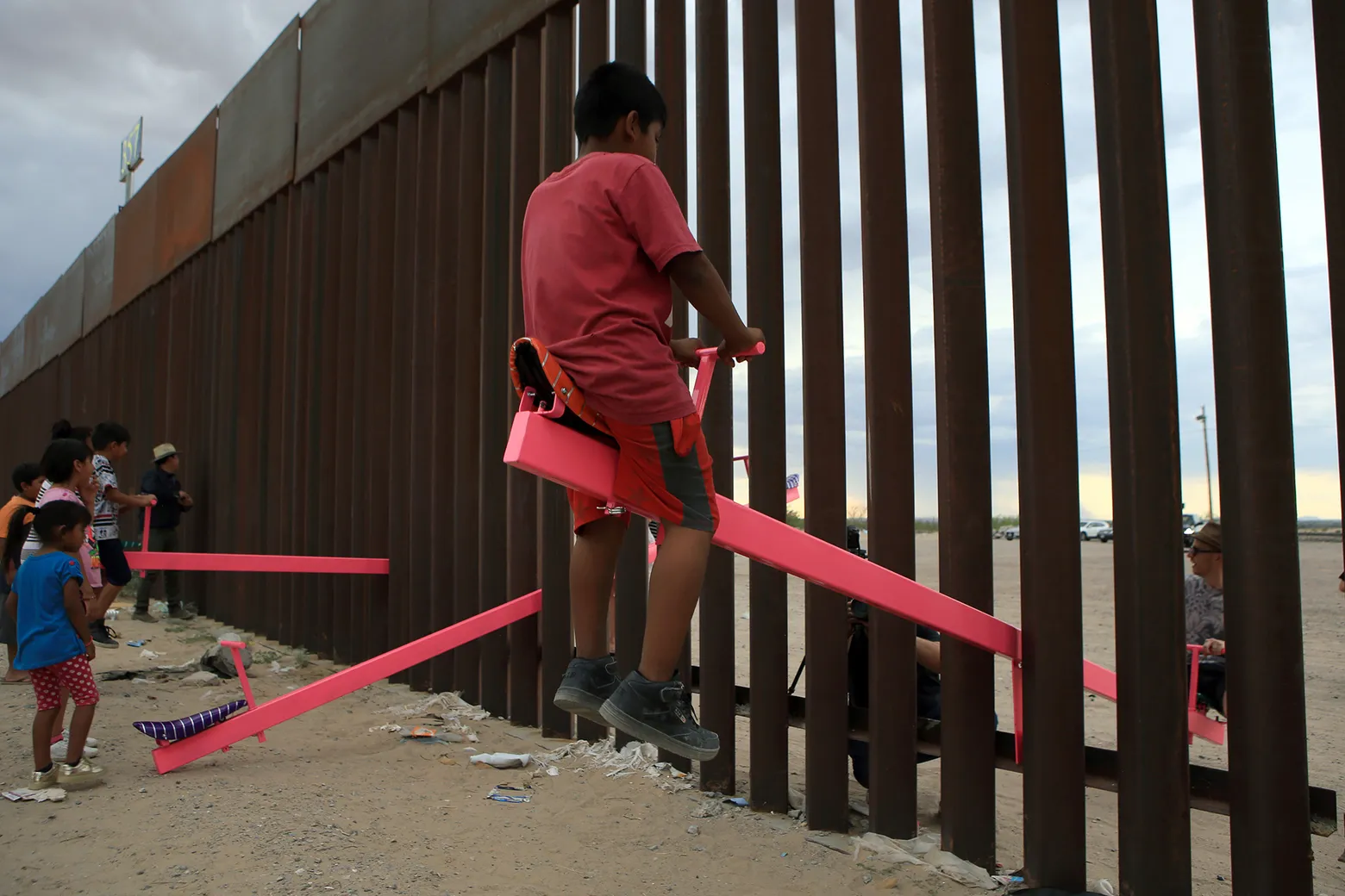 A photograph of a boy in a red shirt and shorts, riding a see-saw that bisects a tall fence. A child on the other side of the fence can be seen crouching down. 